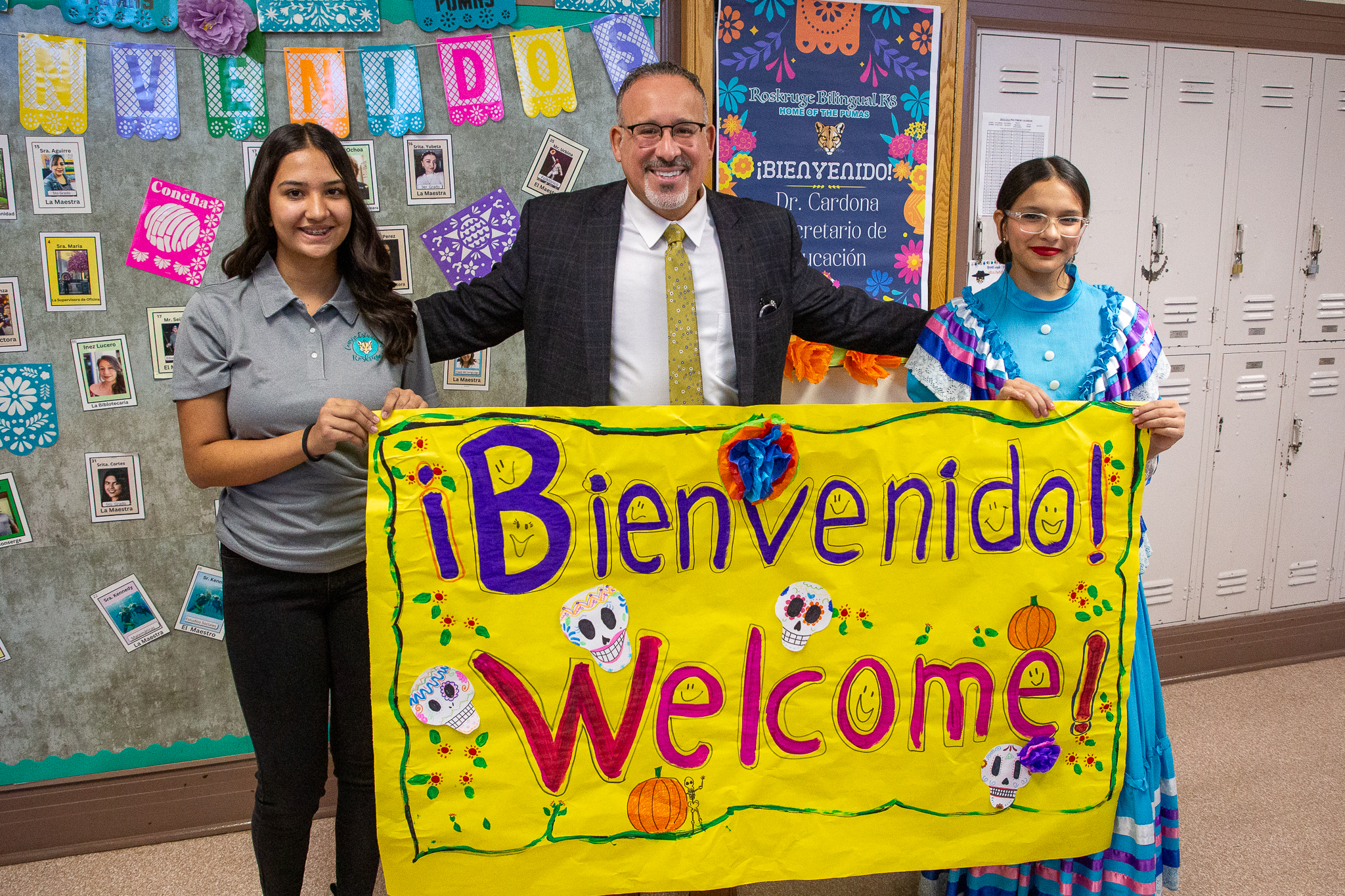 Secretary Cardona and two students pose with a banner reading Bienvenido, Welcome