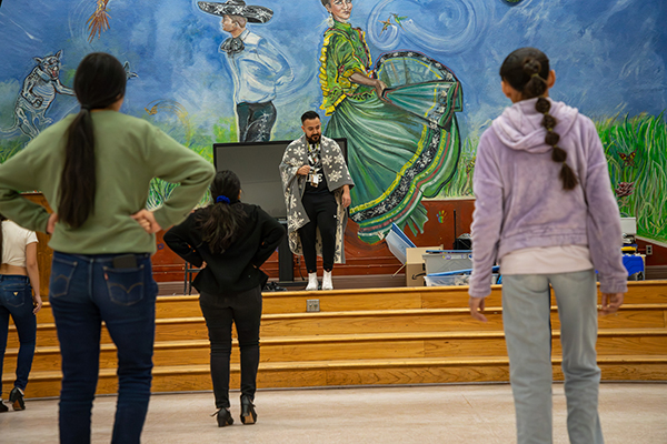 Students stand facing the stage with a folklorico mural