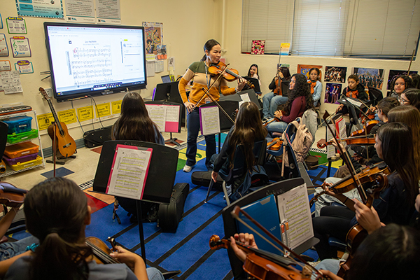 A woman demonstrates how to play the violin to a classroom of students