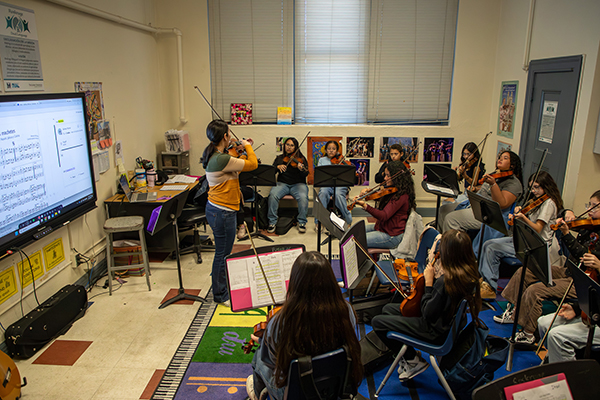 A teacher stands in front of her class playing the violin, as they look on