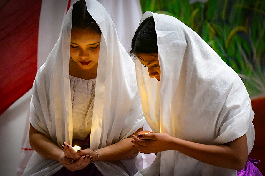 Two girls in white head coverings kneel while holding small candles in their hands