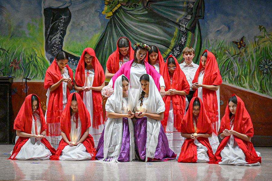 Students in white dresses and red hair coverings gather in a group on stage
