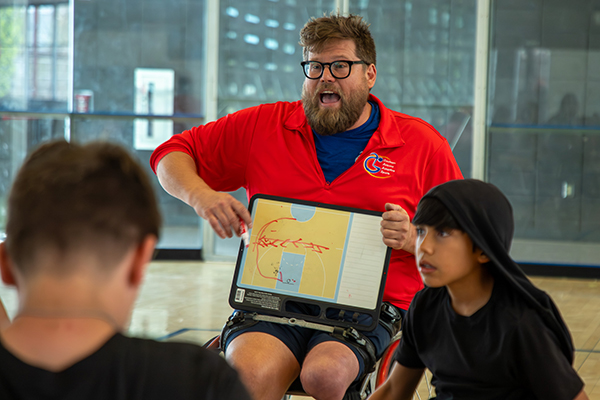 A man sitting in a wheelchair holds up a screen showing a basketball court